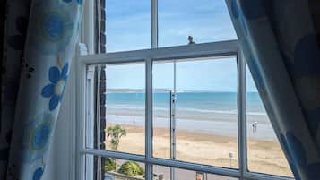 Vista a través de una ventana con cortina floral, que da a una playa con gente y un cielo despejado.