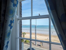 Vista a través de una ventana con cortina floral, que da a una playa con gente y un cielo despejado.