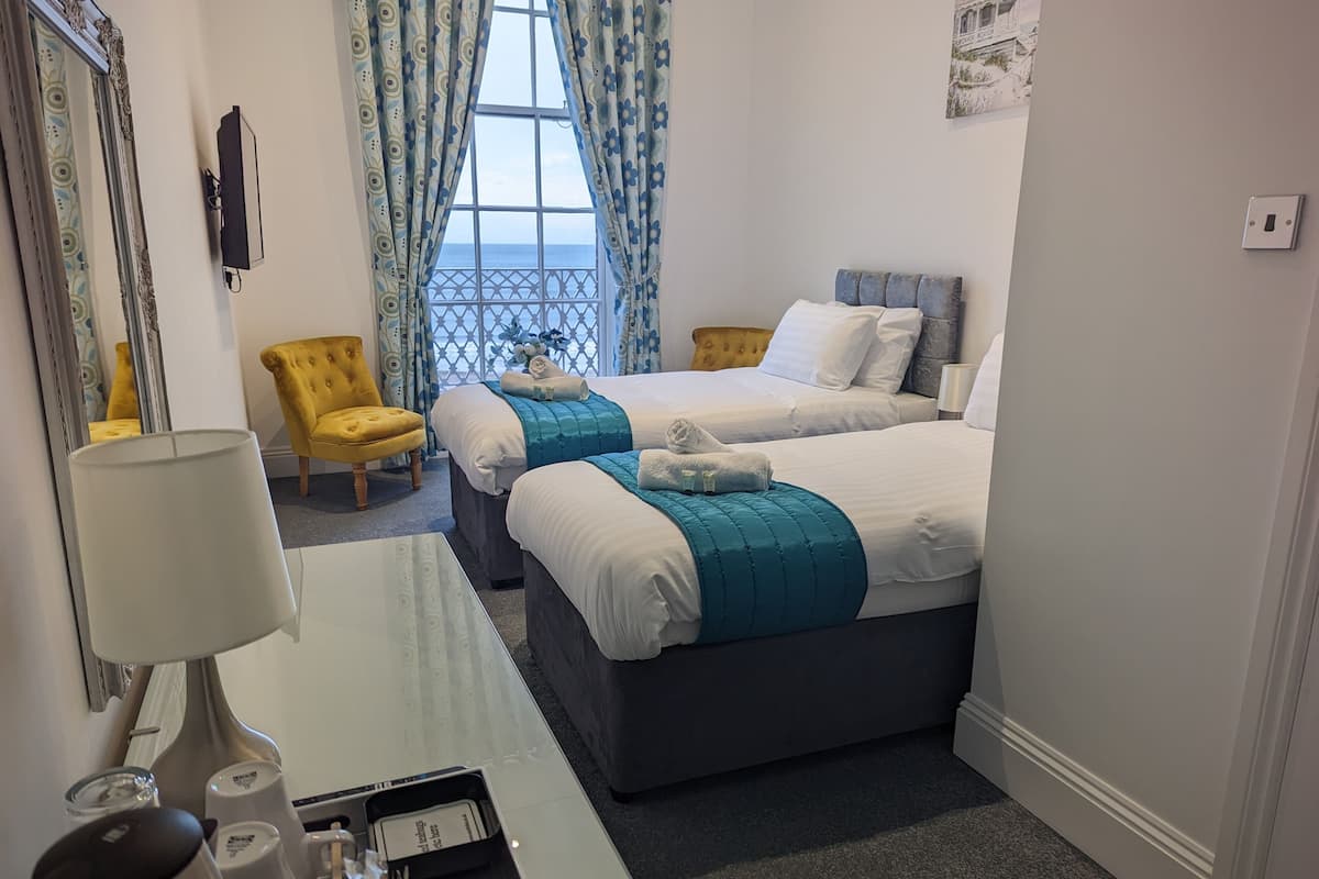 A hotel room with two beds, a desk with a lamp, a chair, and a view of the sea through a window.
