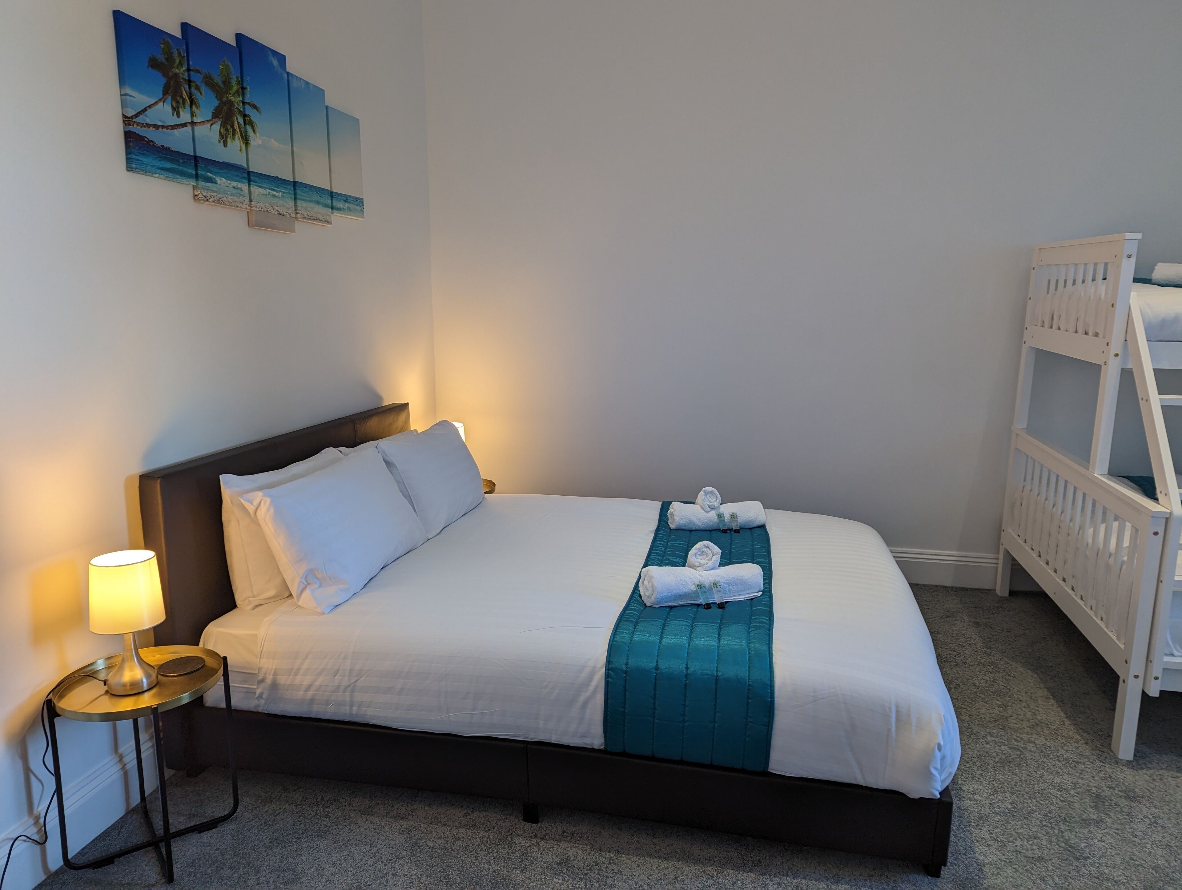 A neatly made bed with white linens and a teal accent, a bedside lamp, and a wall-mounted painting of a beach scene.