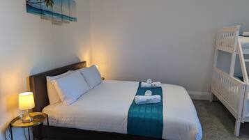 A neatly made bed with white linens and a teal accent, a bedside lamp, and a wall-mounted painting of a beach scene.