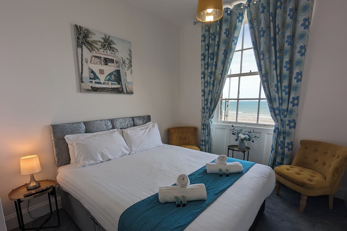 A hotel room with a bed, a nightstand, a chair, a window with curtains, and a painting on the wall.