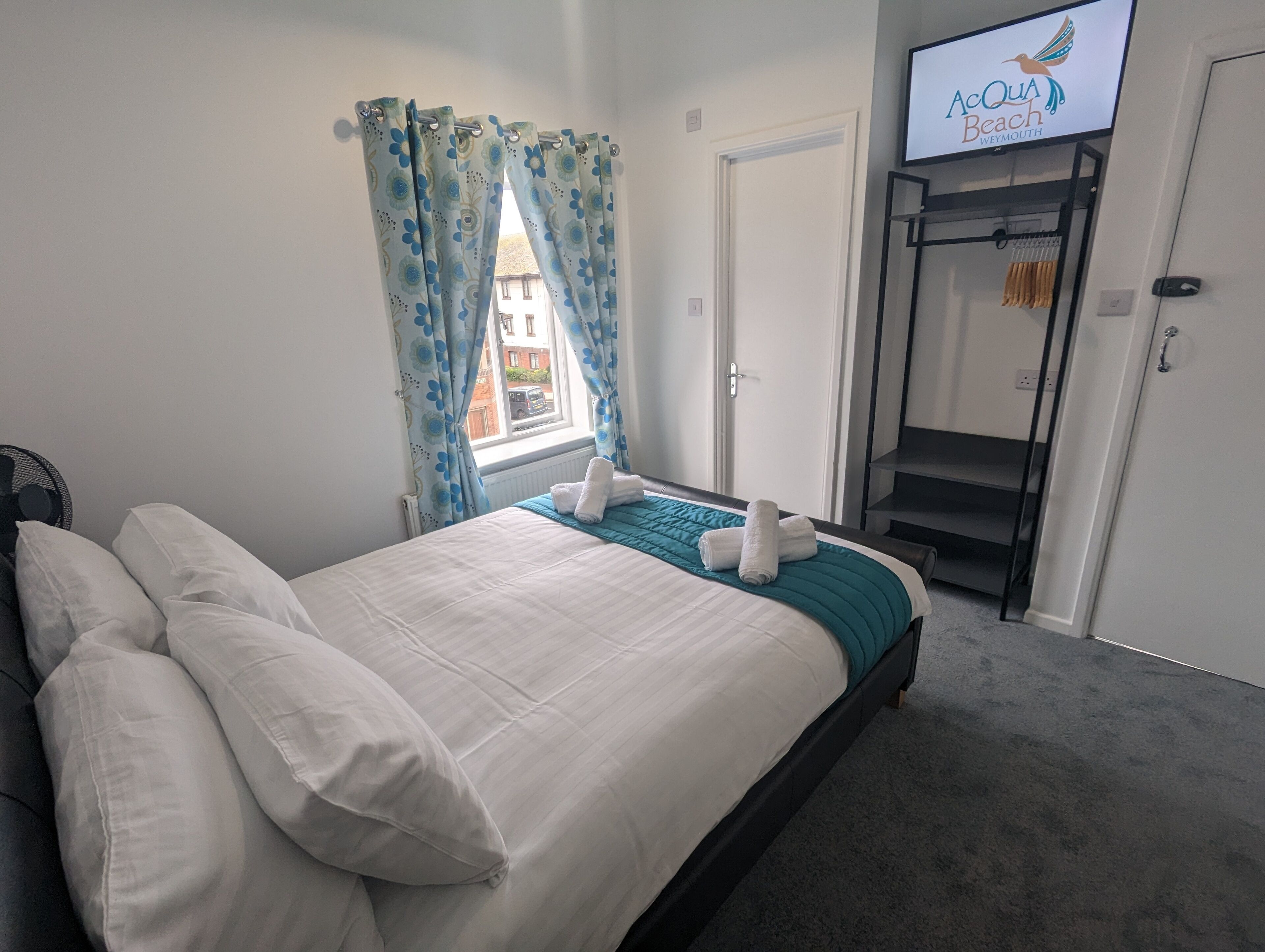 Una cama bien hecha con ropa blanca y un detalle azul aguamarina, una ventana con cortinas florales azules, un televisor mostrando 'Acqua Beach Weymouth', y una escalera con un perchero.
