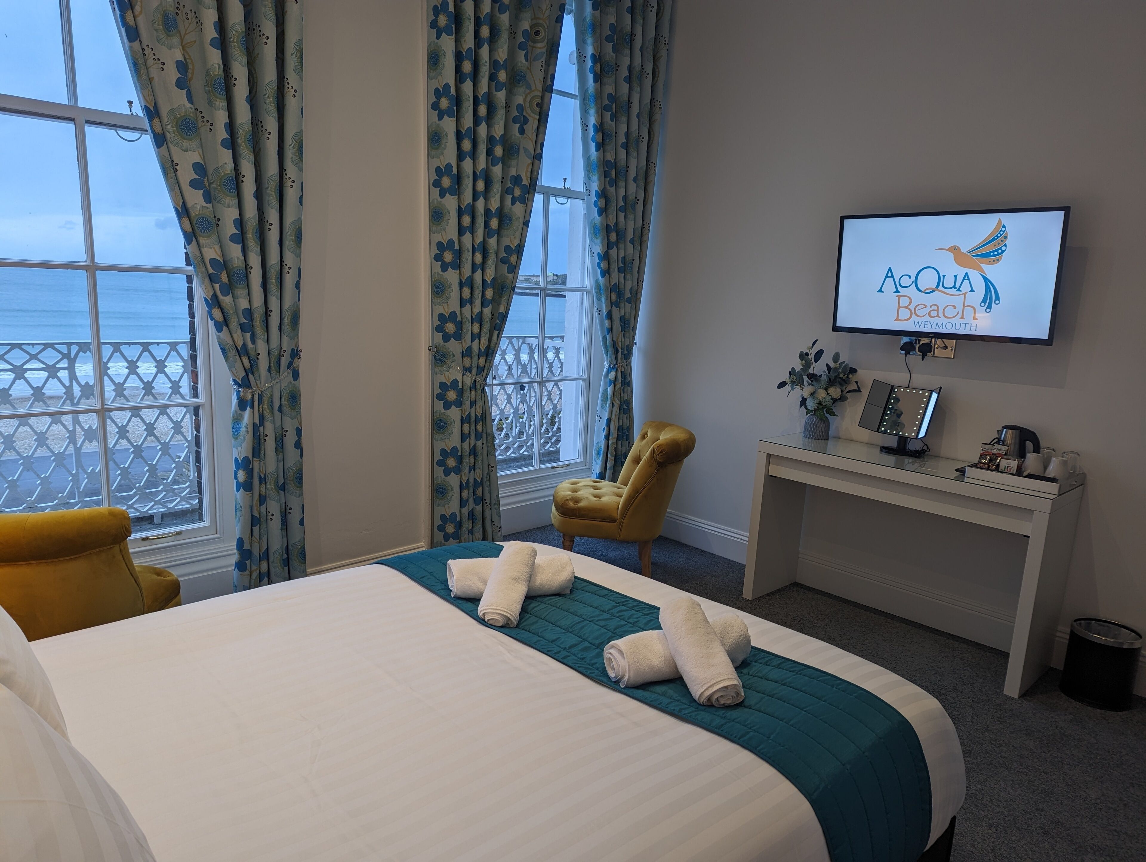 Quarto de hotel com cama, poltrona, secretária e televisão, com vista para o mar.
