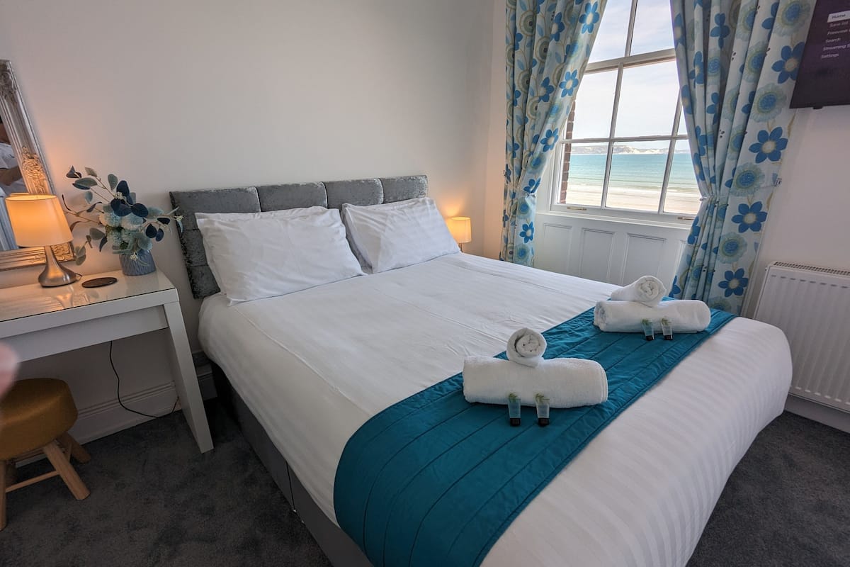 A neatly made bed with white linens and a blue accent, a bedside table with a lamp, and a view of the sea through a window with floral curtains.