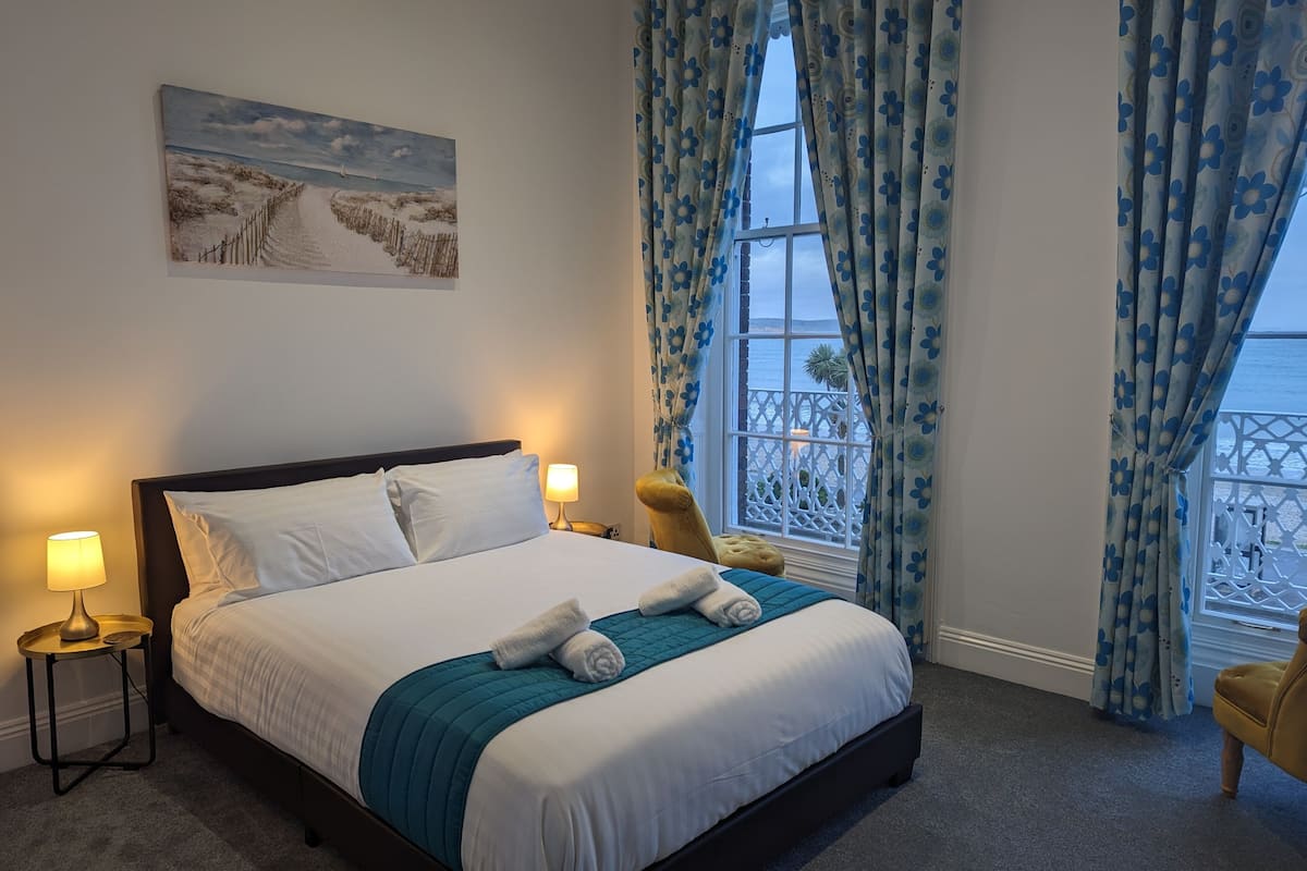 A neatly made bed with white linens and a teal throw, two bedside lamps, a window with blue floral curtains, and a painting of a beach scene on the wall.