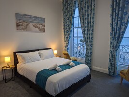 Una cama bien hecha con ropa de cama blanca y un plaid color turquesa, dos lámparas de noche, una ventana con cortinas florales azules y un cuadro de una playa en la pared.