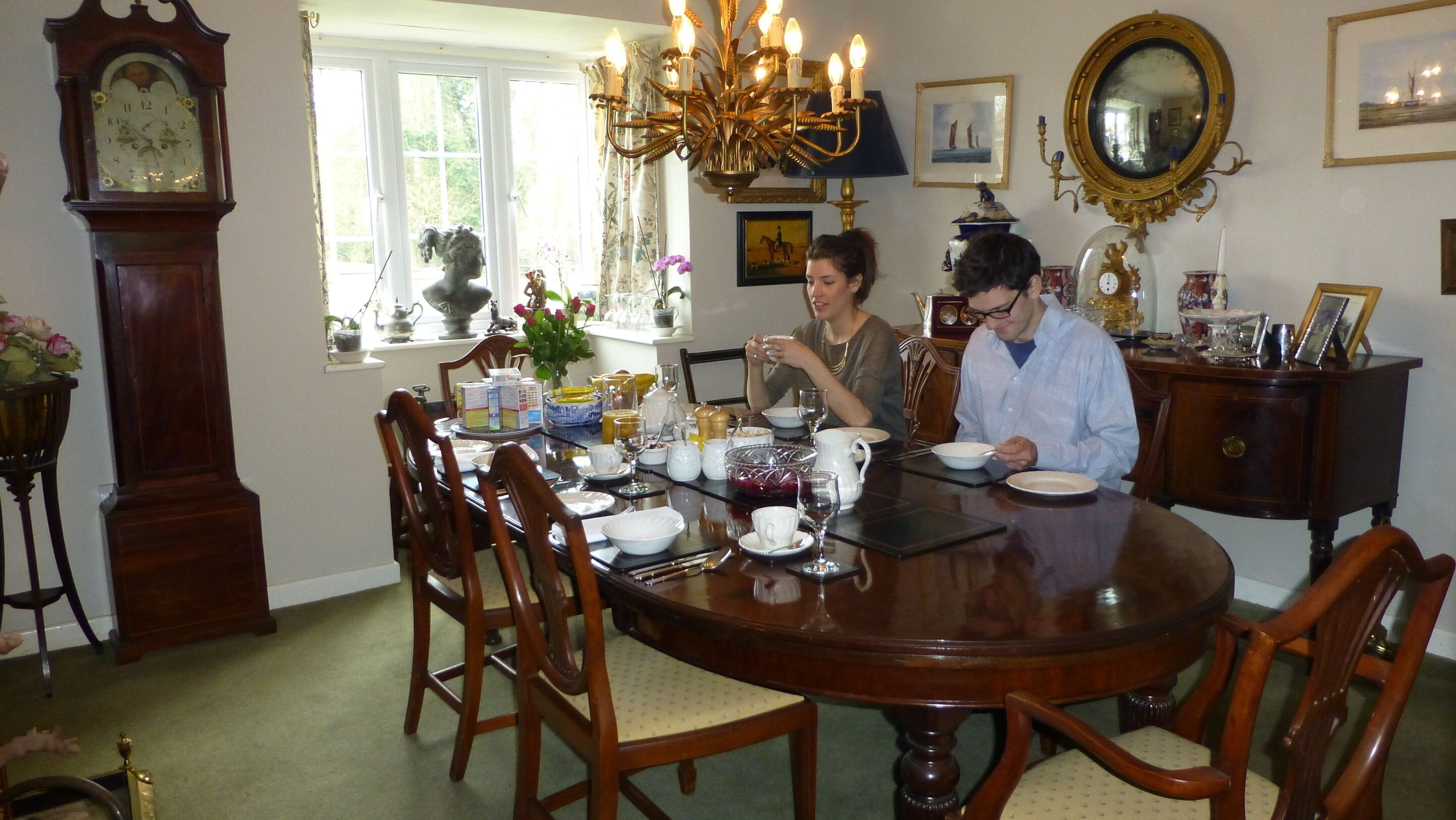 A dining room with a wooden table set for two, surrounded by chairs. There is a large window with curtains, a chandelier, and various decorative items on the walls.