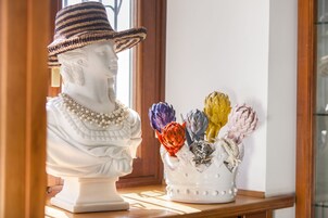 Una escultura de busto con sombrero y collar de perlas sobre una superficie de madera, junto a un jarrón con flores coloridas.