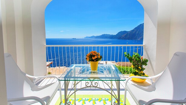 A balcony with a clear view of the sea and mountains, a glass table with a yellow flower vase, and two white chairs. - Hotel Smeraldo (Praiano)