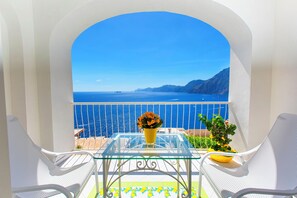 Un balcón con vistas despejadas al mar y a las montañas, una mesa de vidrio con un florero amarillo y dos sillas blancas.