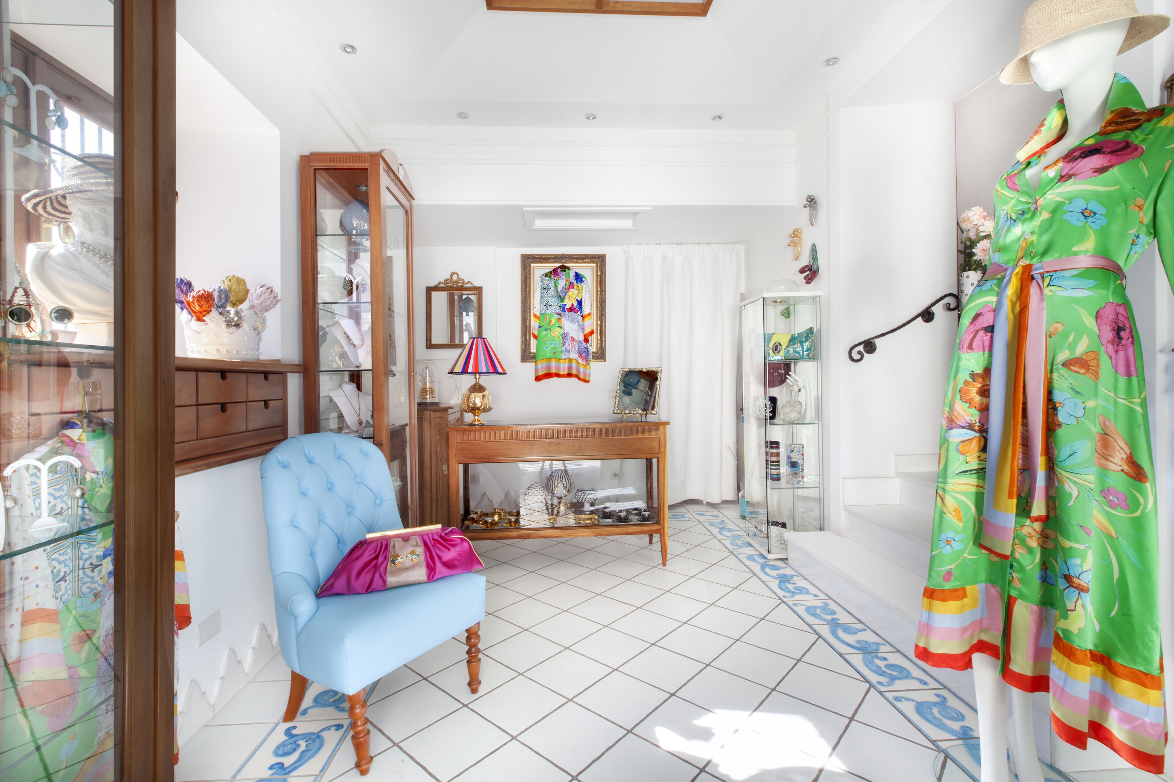 A room with a blue armchair, a wooden console table, a glass cabinet, and a colorful floral dress on a mannequin.
