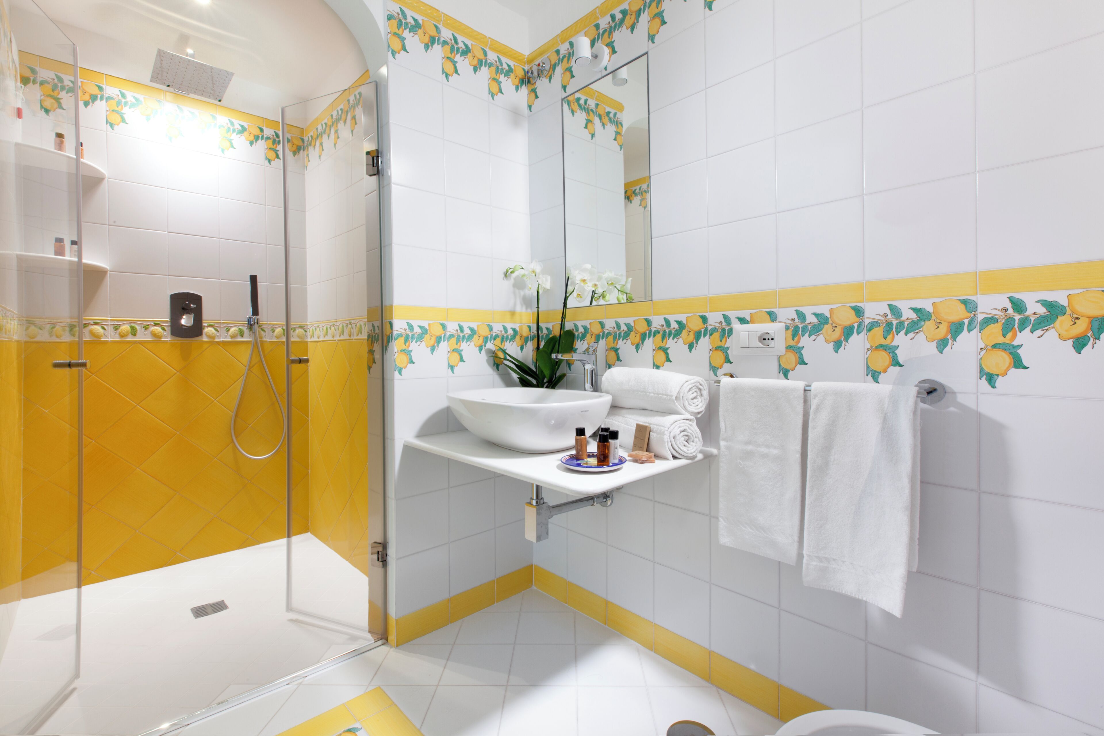 A bathroom with a glass-enclosed shower, a white sink with a mirror above it, and yellow and white tiles with a floral pattern.