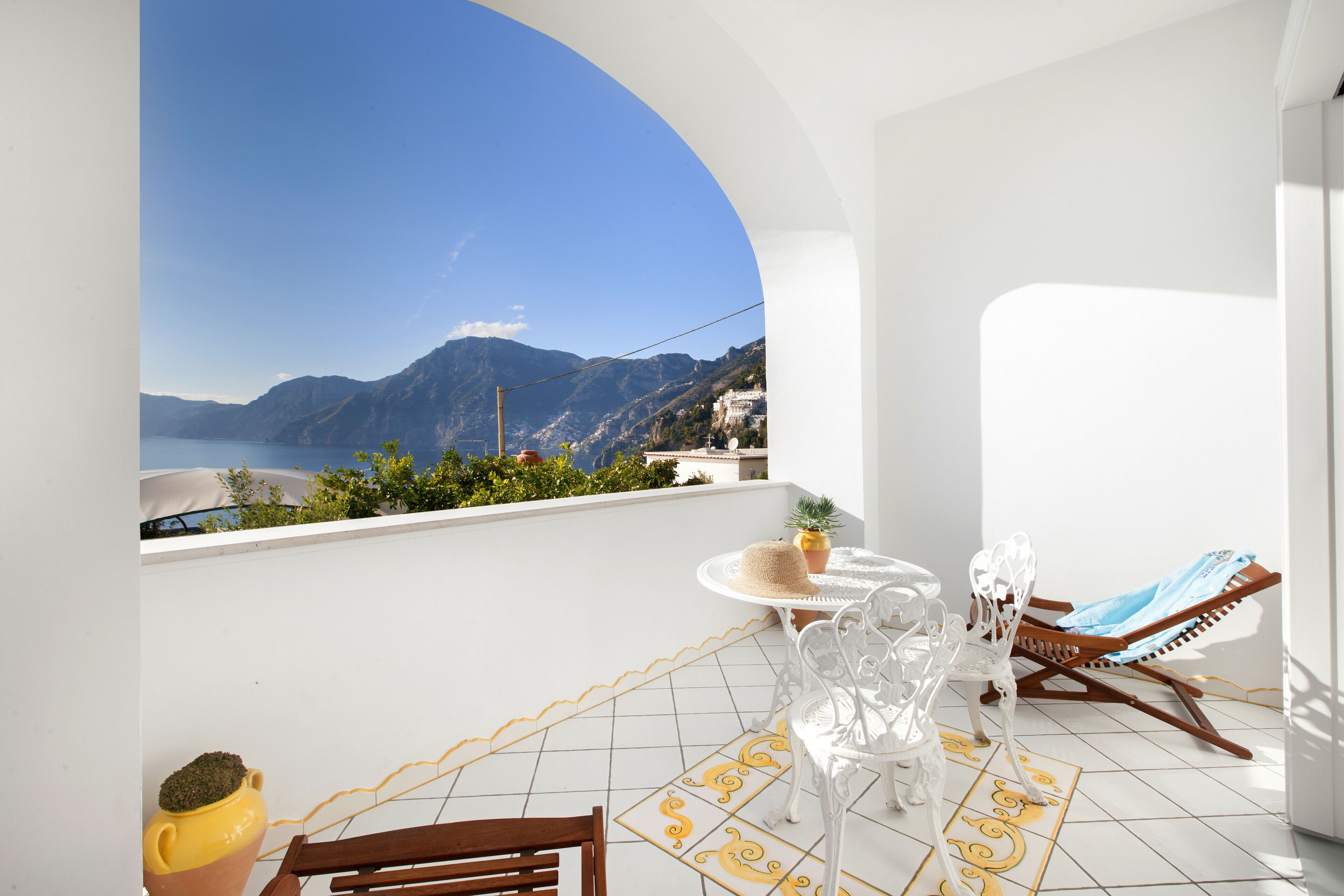 A balcony with a white tiled floor, a round table with chairs, a wooden chair, a sun lounger, and a view of mountains and a lake.