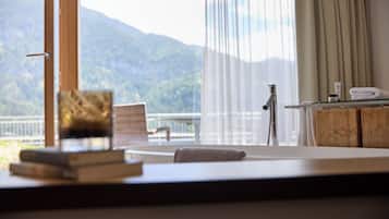 Ein Zimmer mit Blick auf Berge, ein großes Fenster mit Vorhängen, ein Sessel, ein kleiner Tisch mit einem Buch und einem Handtuch sowie eine Holzkommode mit Toilettenartikeln.