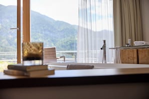 A room with a view of mountains, a large window with curtains, a chair, a small table with a book and a towel, and a wooden cabinet with toiletries.