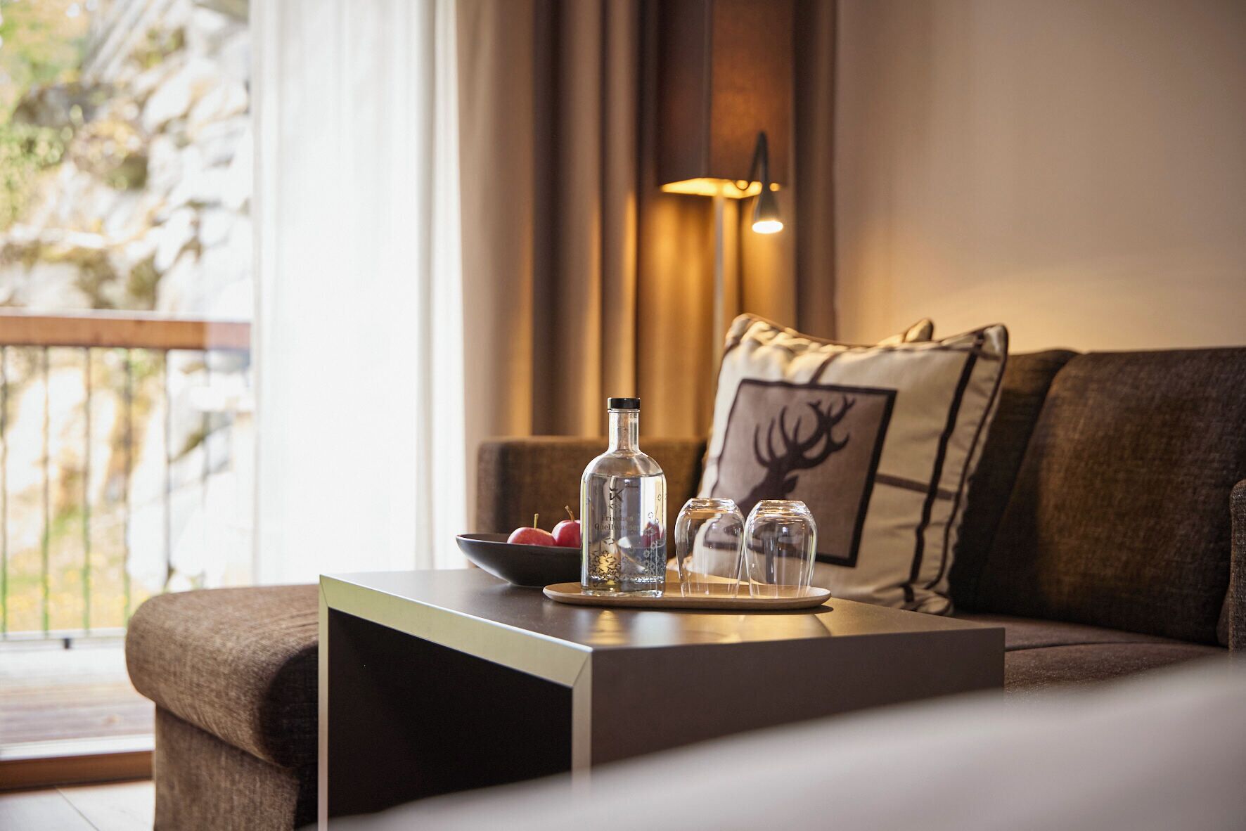 A modern living room with a sofa, a side table with a bottle and glasses, a bowl of fruit, and a decorative pillow with a deer design.