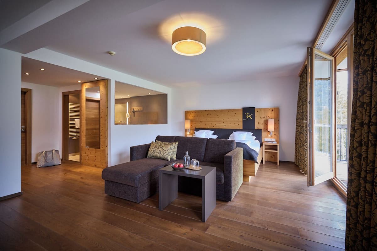 Una habitación de hotel moderna con suelo de madera, un sofá, una cama, una mesita de noche y un ventanal grande.
