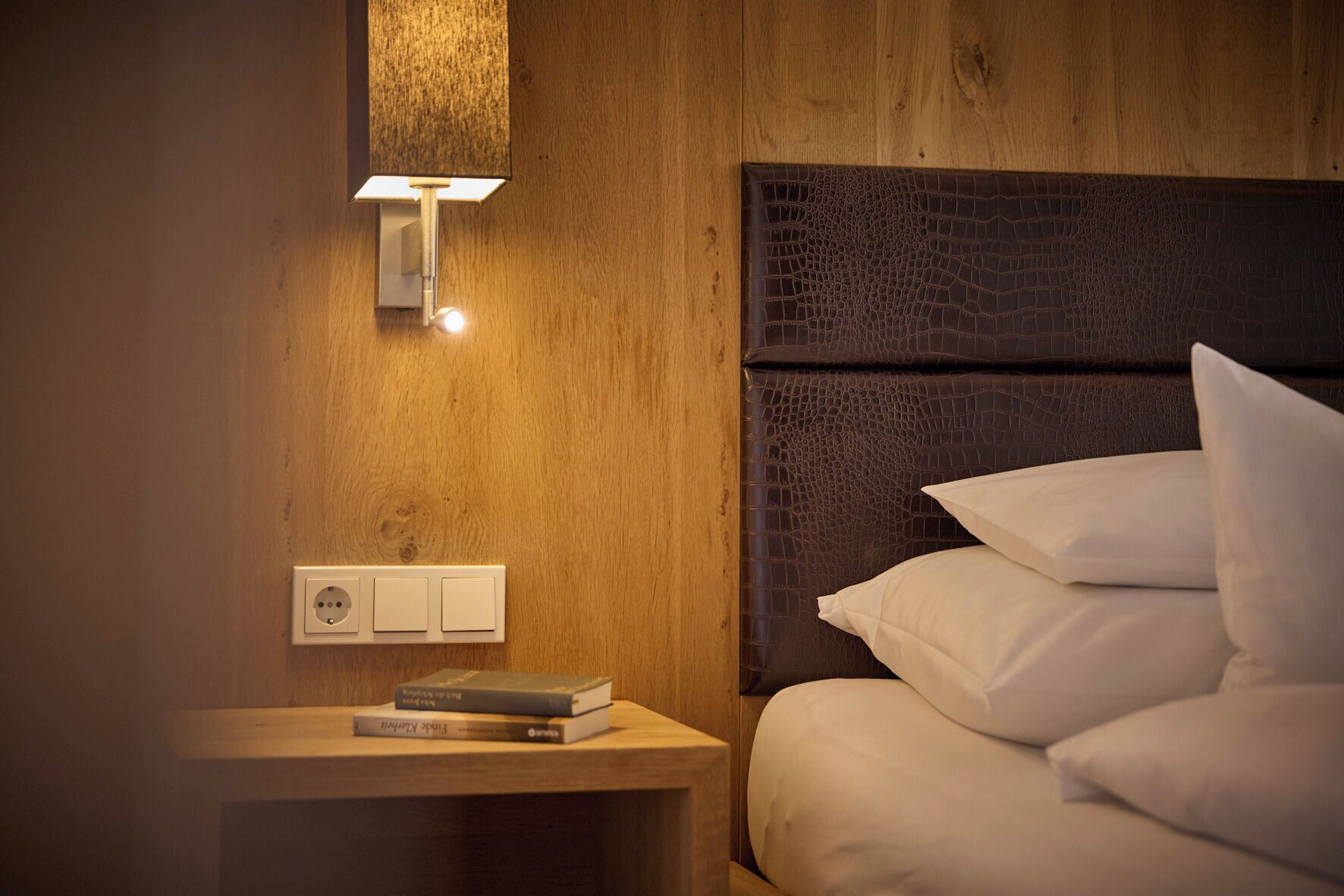 A wooden wall with a textured headboard, a bedside lamp, a power outlet, and a stack of books.