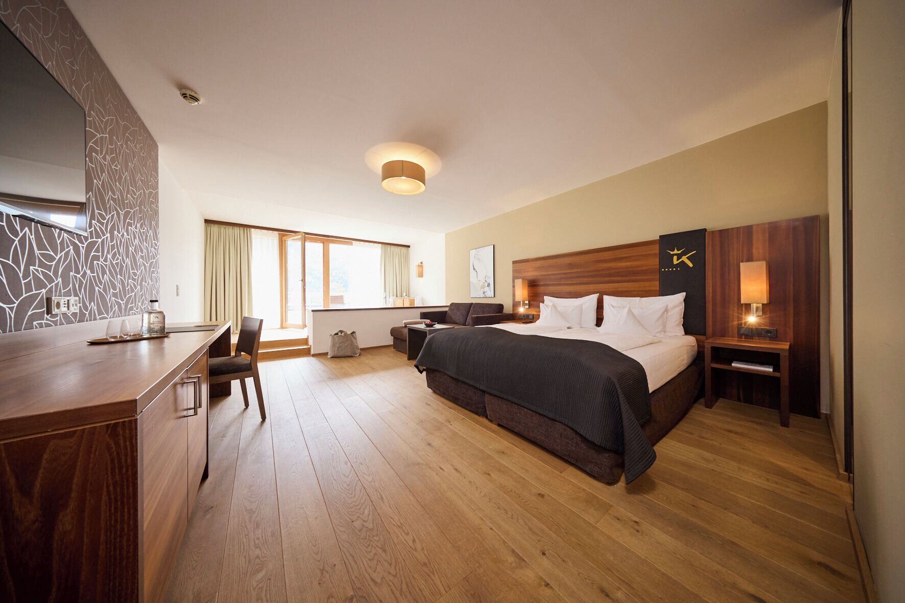 A hotel room with a bed, bedside tables, a desk, a chair, a window, and a wall with a decorative pattern.