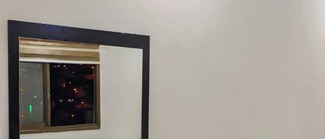 Una habitación de hotel con una cama, una mesita de noche con un control remoto, un espejo grande que refleja la vista por la ventana y una lámpara en el techo.