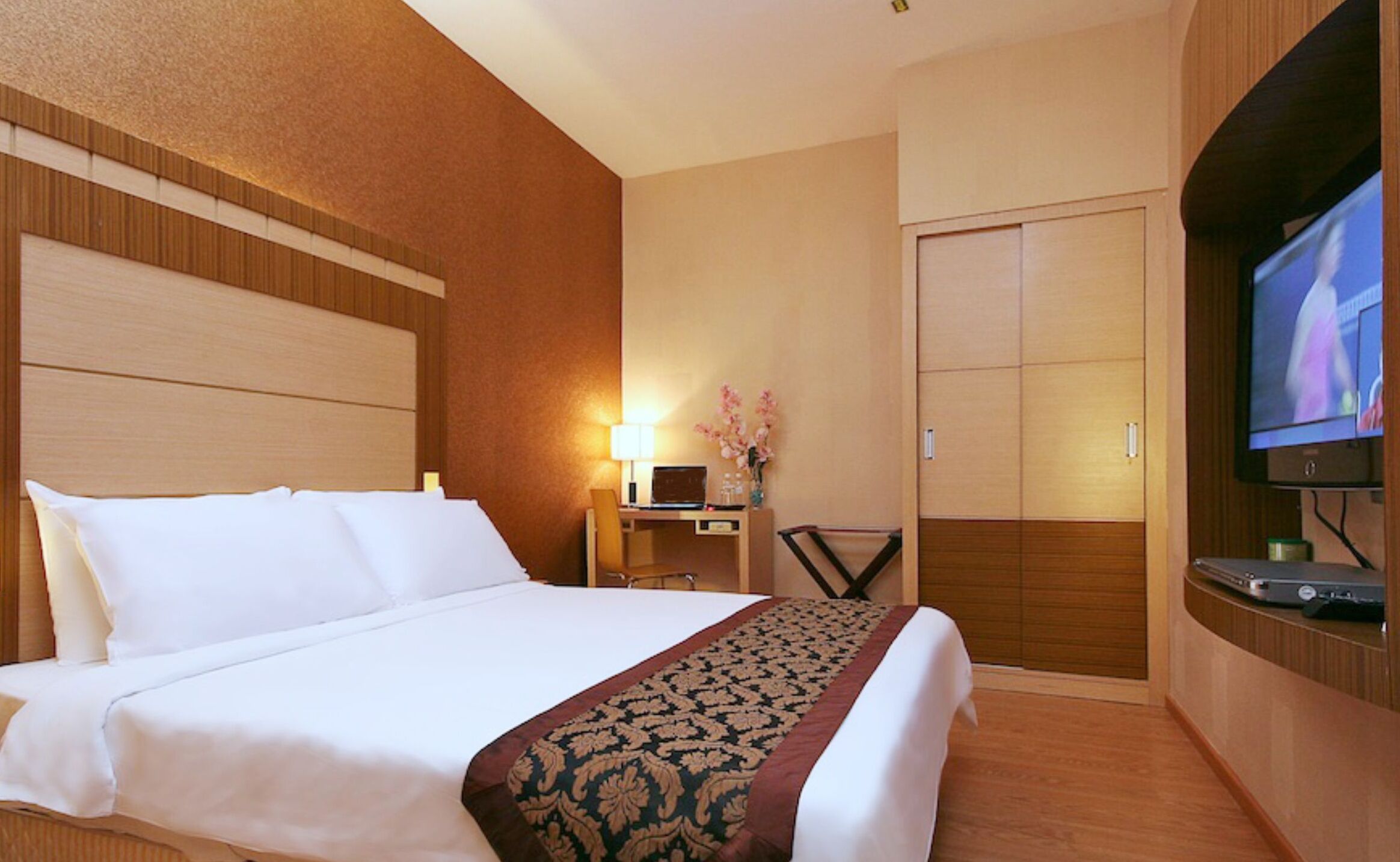 A hotel room with a bed, a desk with a lamp, a TV, and a wardrobe.