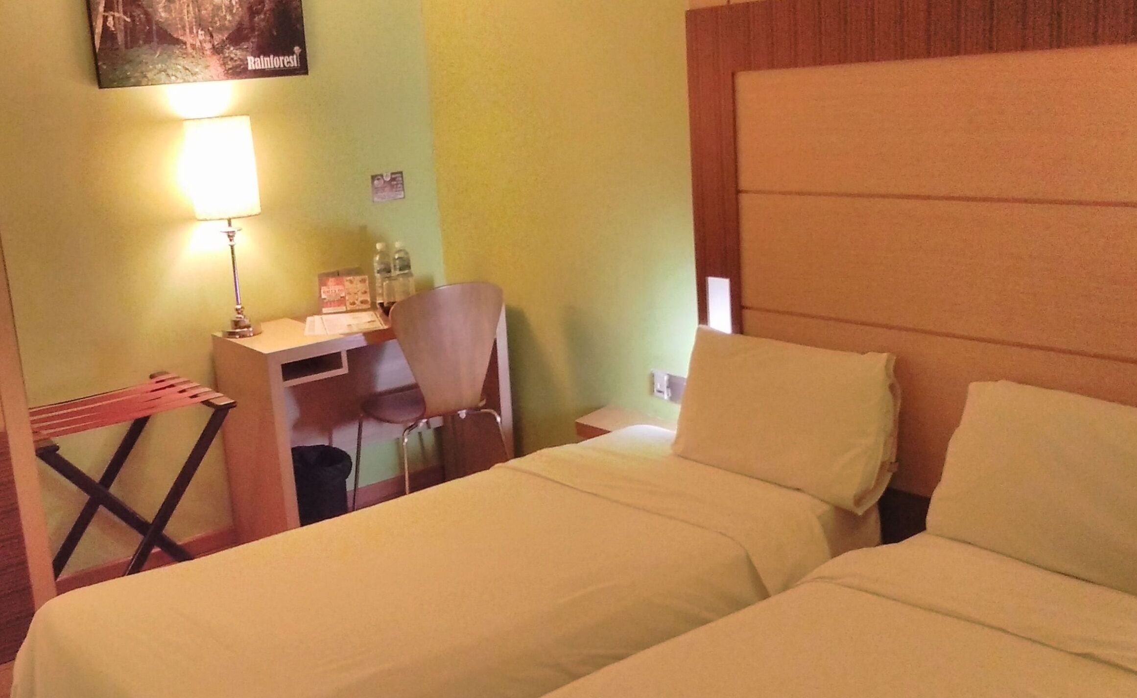 A hotel room with two beds, a desk with a chair, a lamp, and a small table with a bottle and a menu.