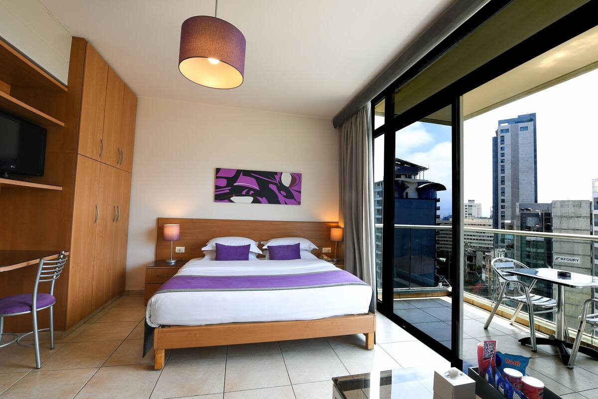 A hotel room with a bed, a desk, a chair, a TV, a painting, and a balcony with a view of the city.