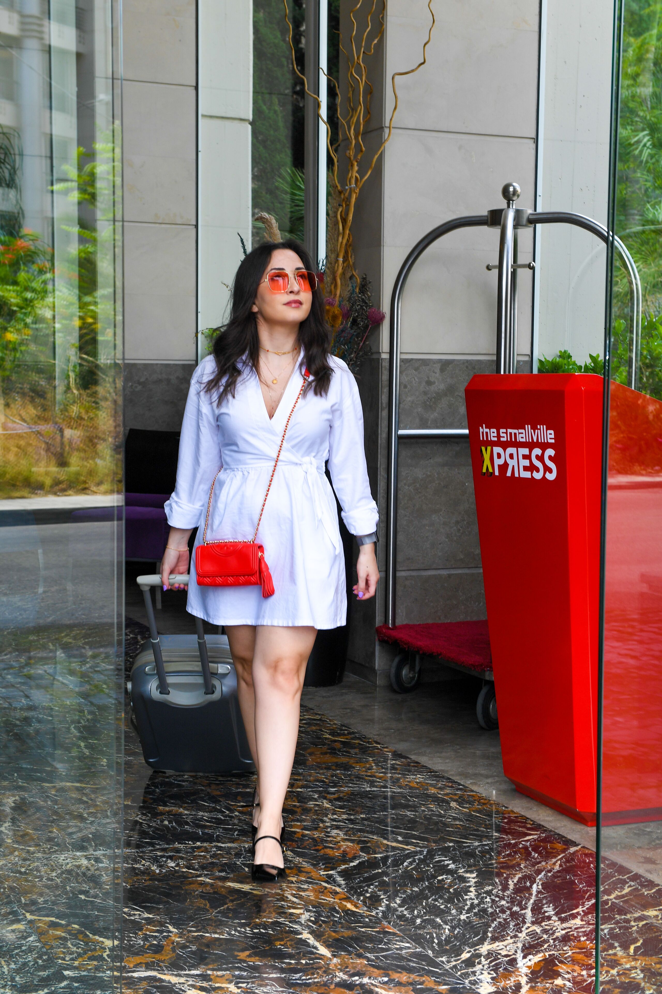 Una mujer con un vestido blanco y un bolso rojo camina hacia un mostrador de recepción rojo con la leyenda "the smallville XPRESS".