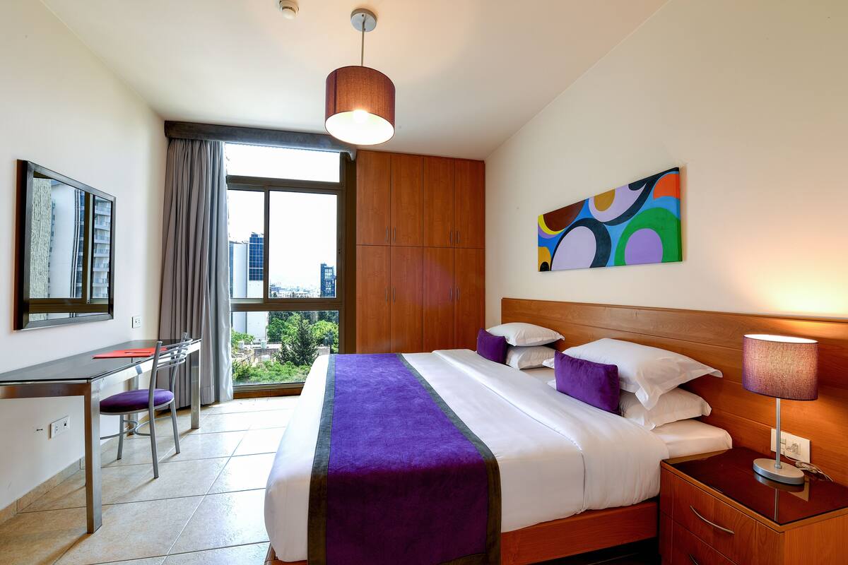A hotel room with a bed, a desk, a chair, a window with a view, and a painting on the wall.