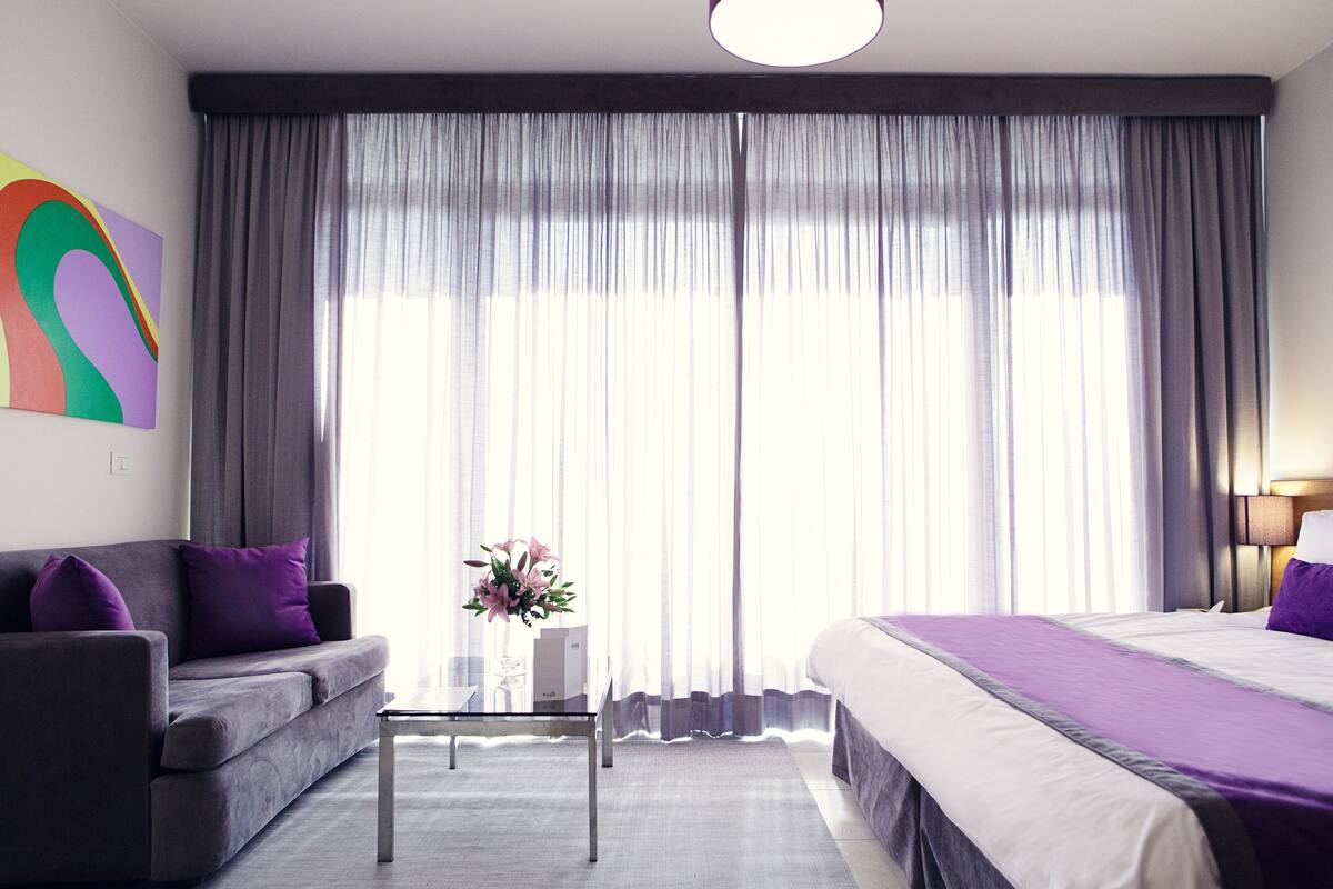 A hotel room with a bed, a sofa, a small table, and a large window with curtains.