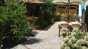 A patio area with outdoor seating and umbrellas, surrounded by greenery and a building with a red roof. - Hotel Heideparadies (Soltau)