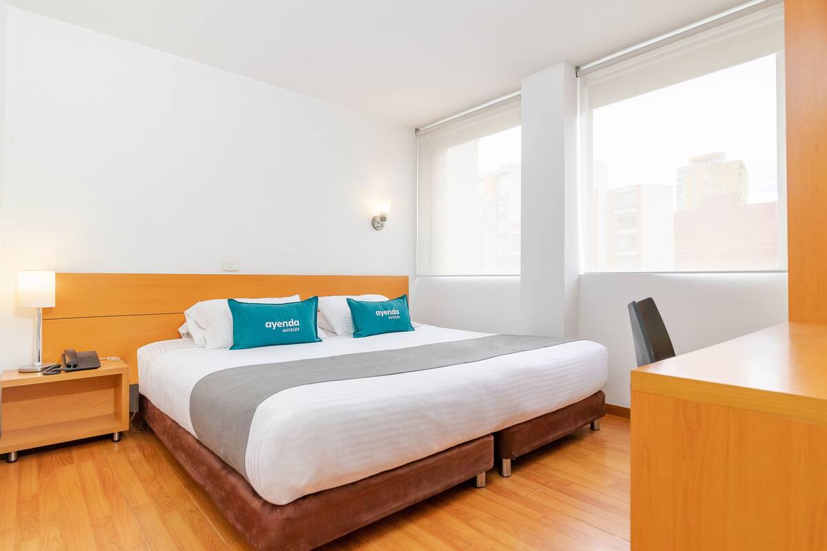 A hotel room with a bed, a desk, a chair, a nightstand with a phone, and a window with blinds.