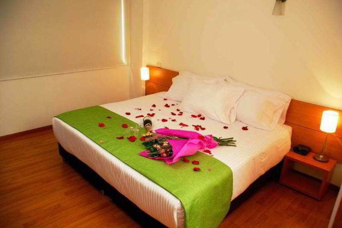 A neatly made bed with a green bedspread, white pillows, and rose petals scattered on the bed.