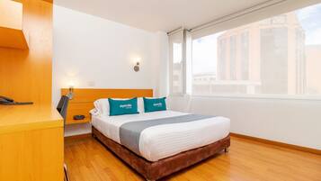 Habitació d'hotel amb escriptori de fusta, llit amb capçalera marró i vista d'un edifici per la finestra.