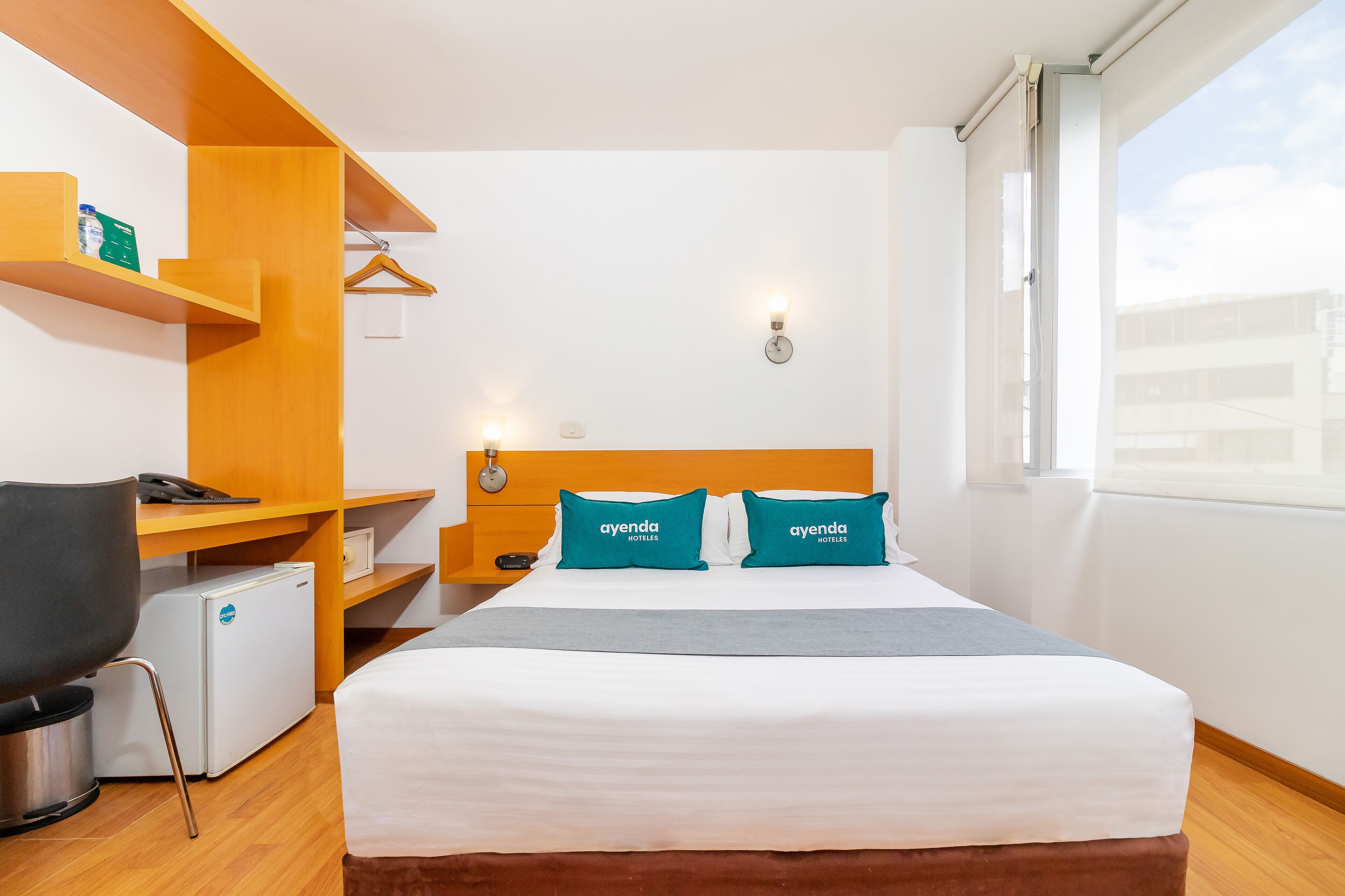 Habitación de hotel con cama, escritorio, silla, mini refrigerador y ventana con vista a edificios.