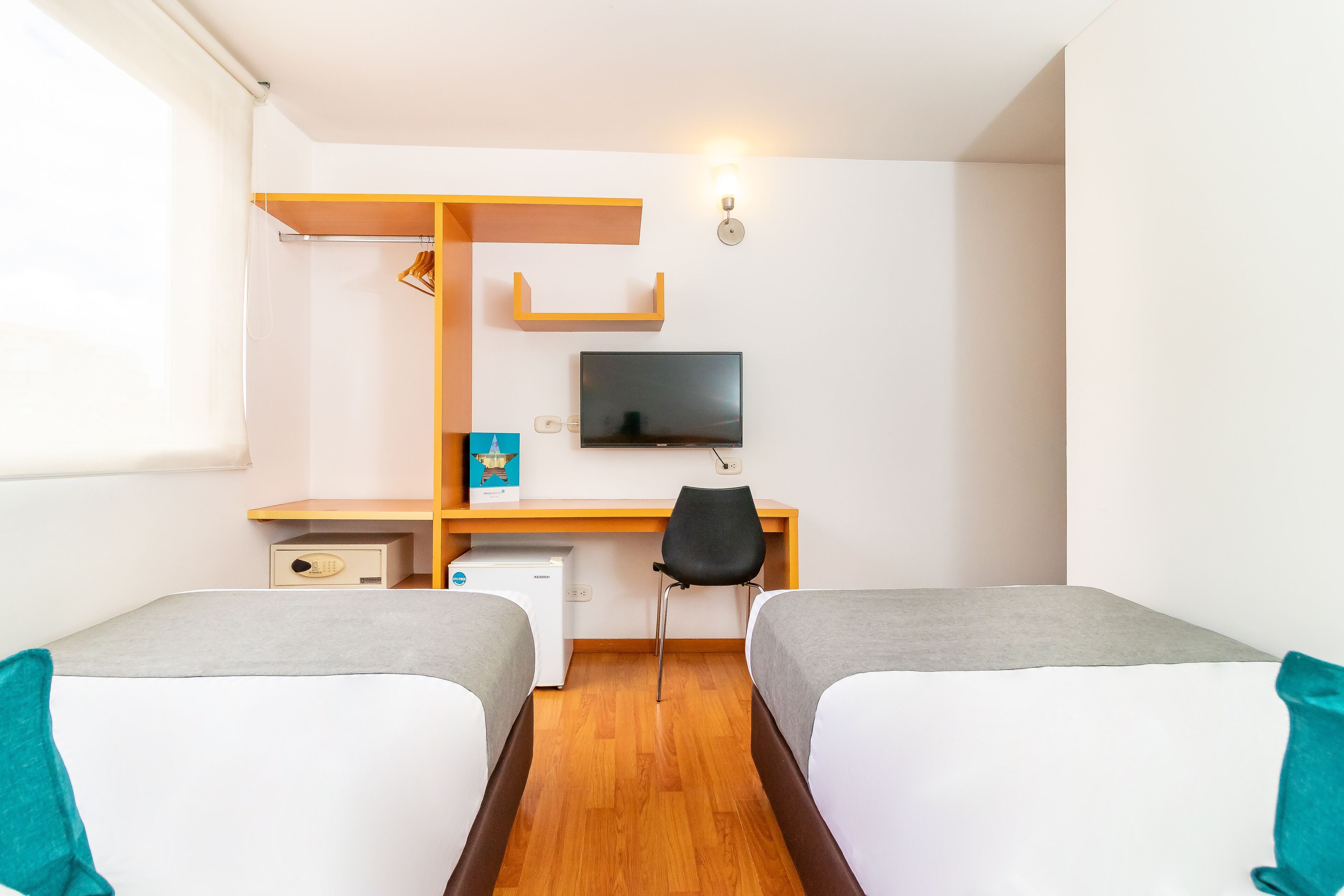 A hotel room with two single beds, a desk with a chair, a TV mounted on the wall, and a small refrigerator.
