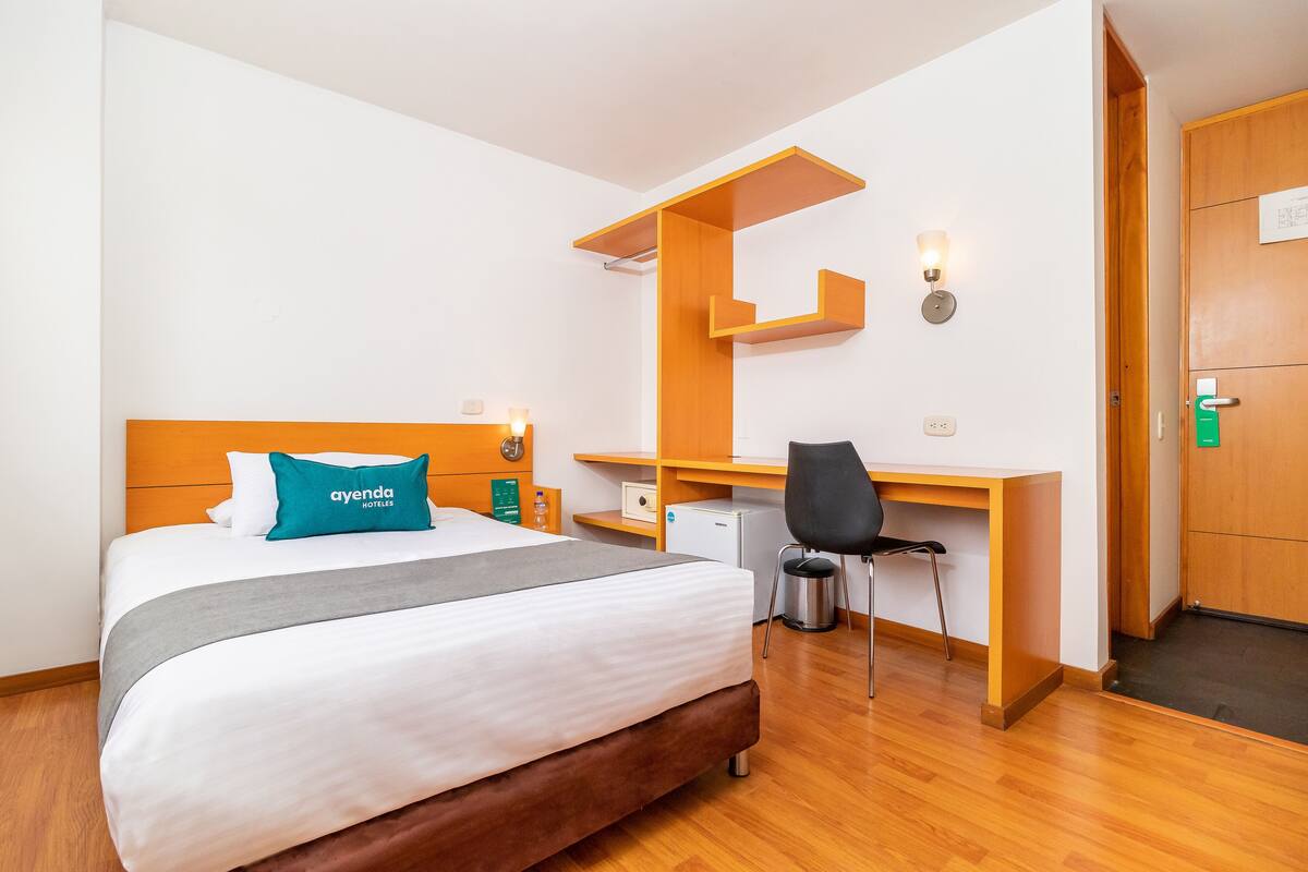 A hotel room with a bed, a desk, a chair, a trash bin, and a wooden door.