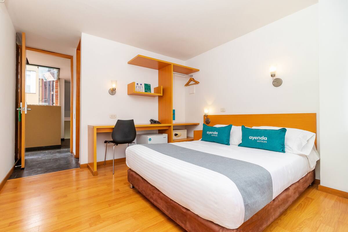 A hotel room with a bed, desk, chair, and a small shelf with toiletries.