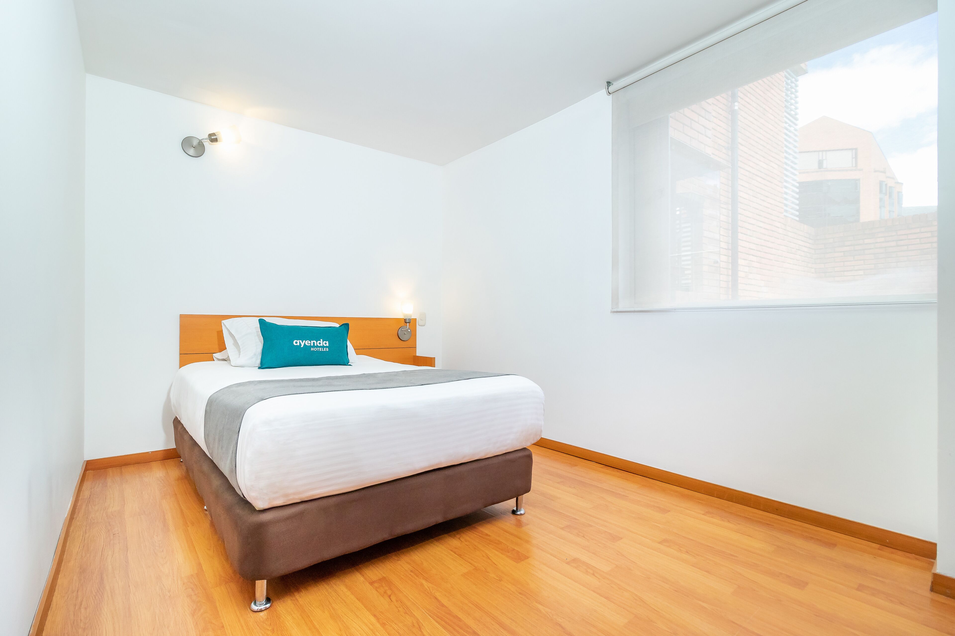 Une chambre d’hôtel moderne avec un lit simple, une tête de lit en bois, une table de chevet, une lampe fixée au mur et une grande fenêtre donnant sur des immeubles.