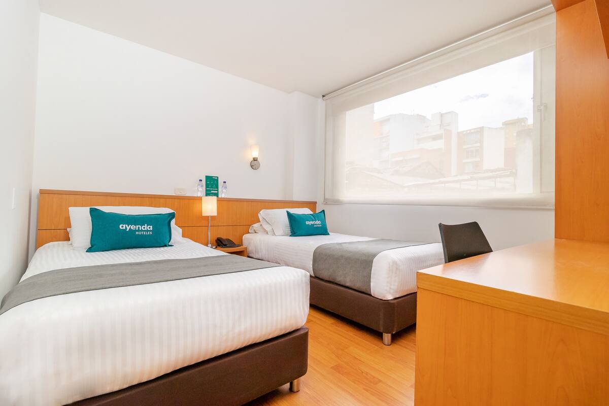 A hotel room with two beds, a desk, a nightstand, and a window with a view of buildings.