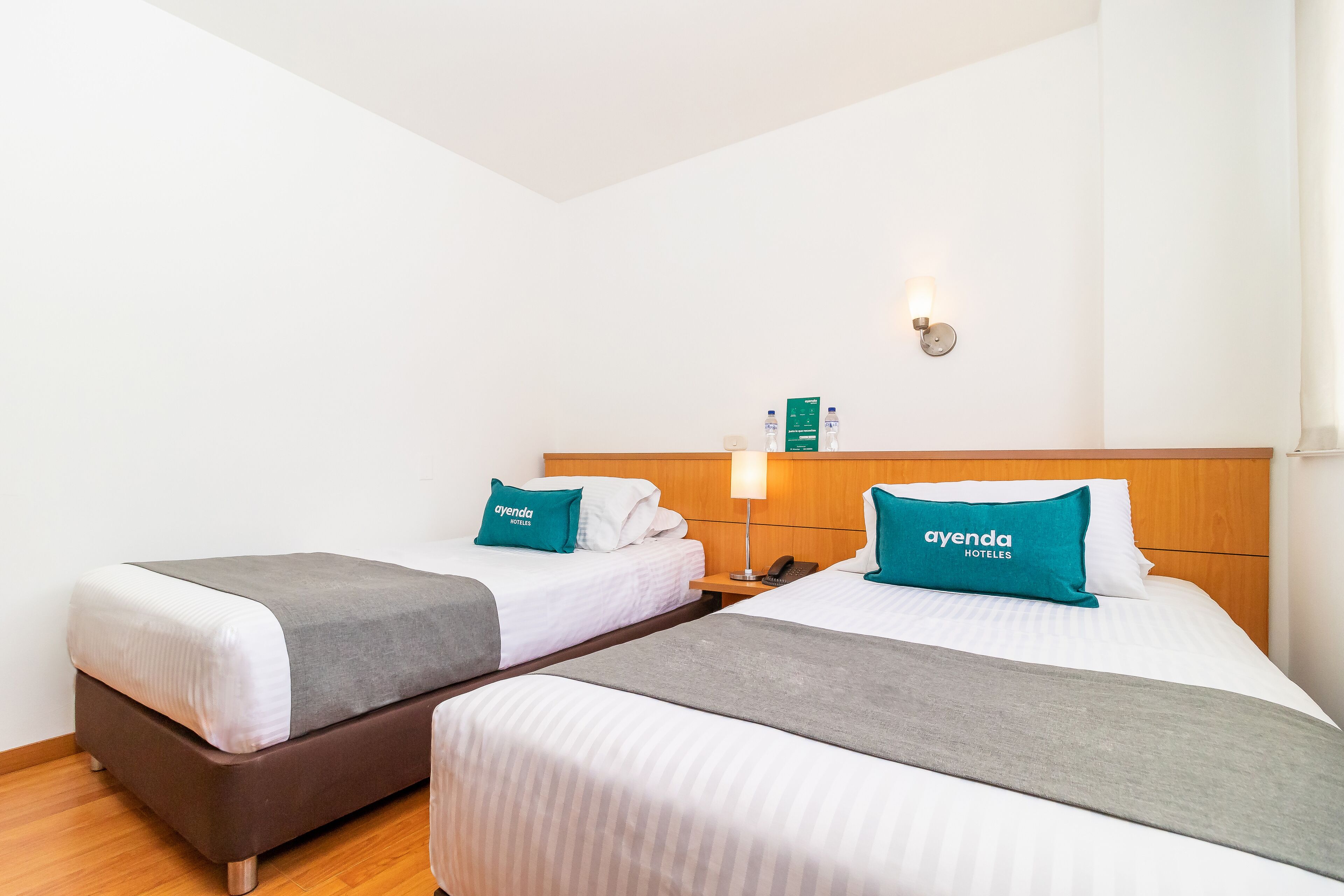 A hotel room with two beds, each with a grey blanket and a teal pillow labeled 'ayenda hoteles'. There is a nightstand with a lamp and a phone between the beds.
