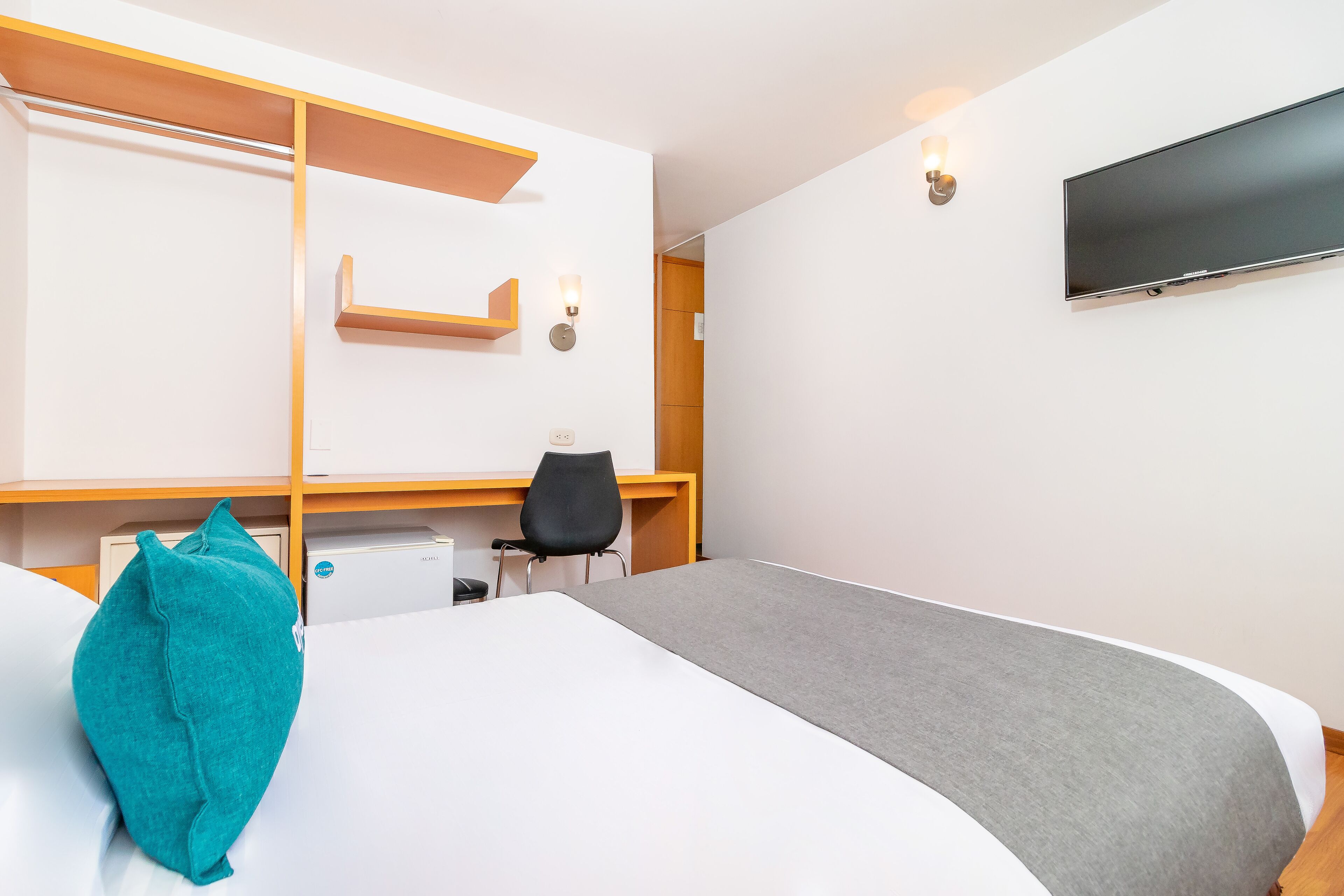 A hotel room with a bed, a desk, a chair, a TV, and a wall-mounted shelf.
