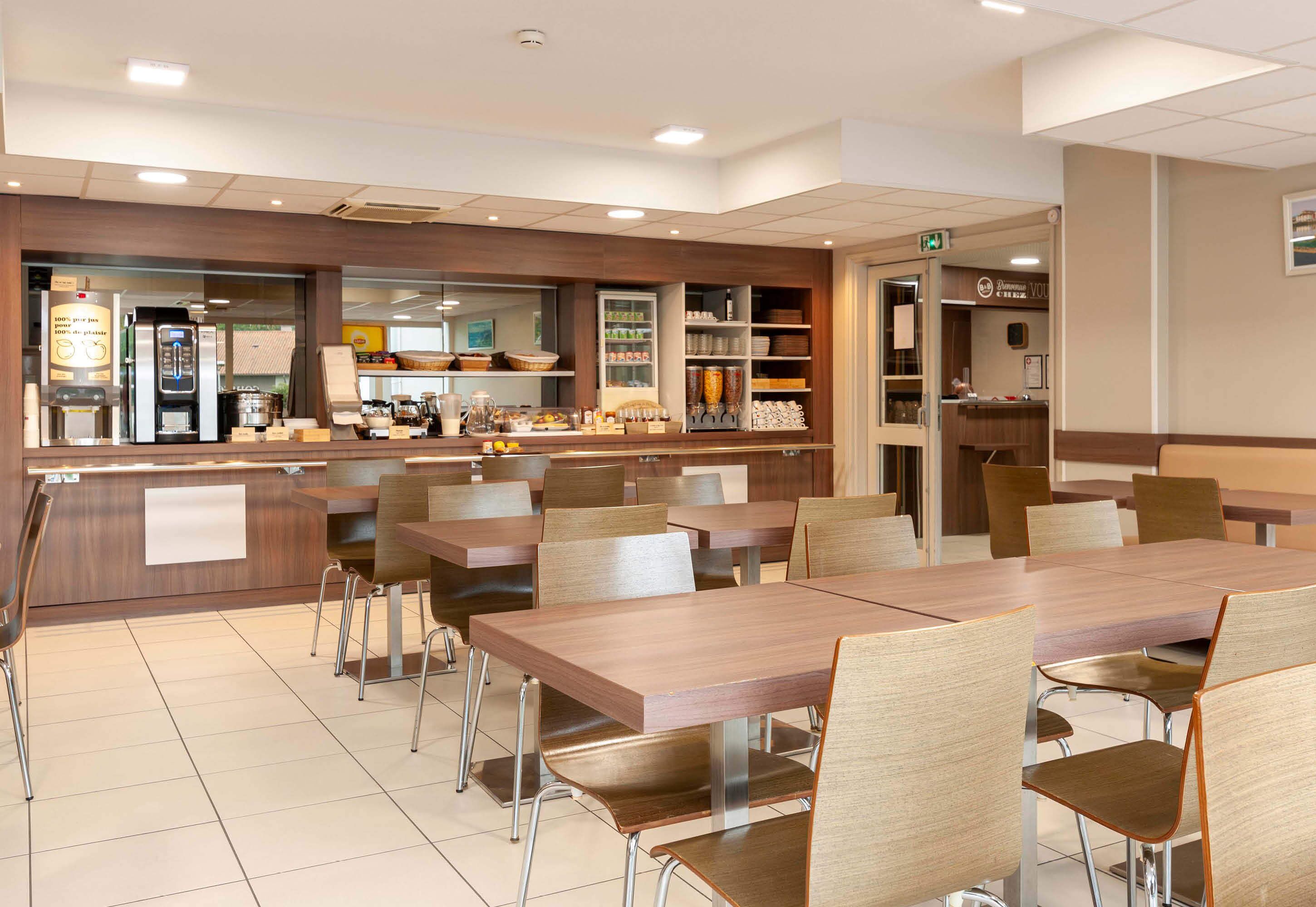 A modern cafeteria with wooden tables and chairs, a self-service beverage station, and a variety of food items on display.