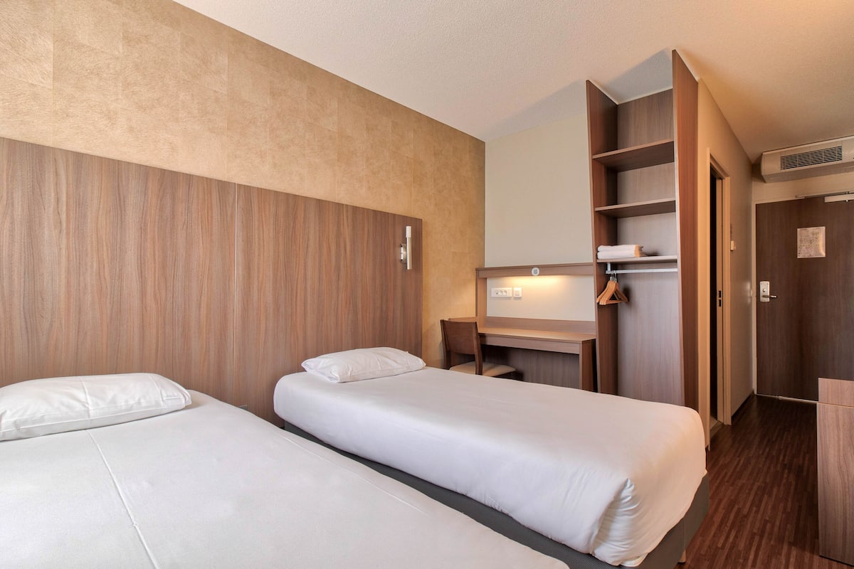 Ett hotellrum med två separata sängar, en sänggavel i trä, ett skrivbord med en stol och en garderob.