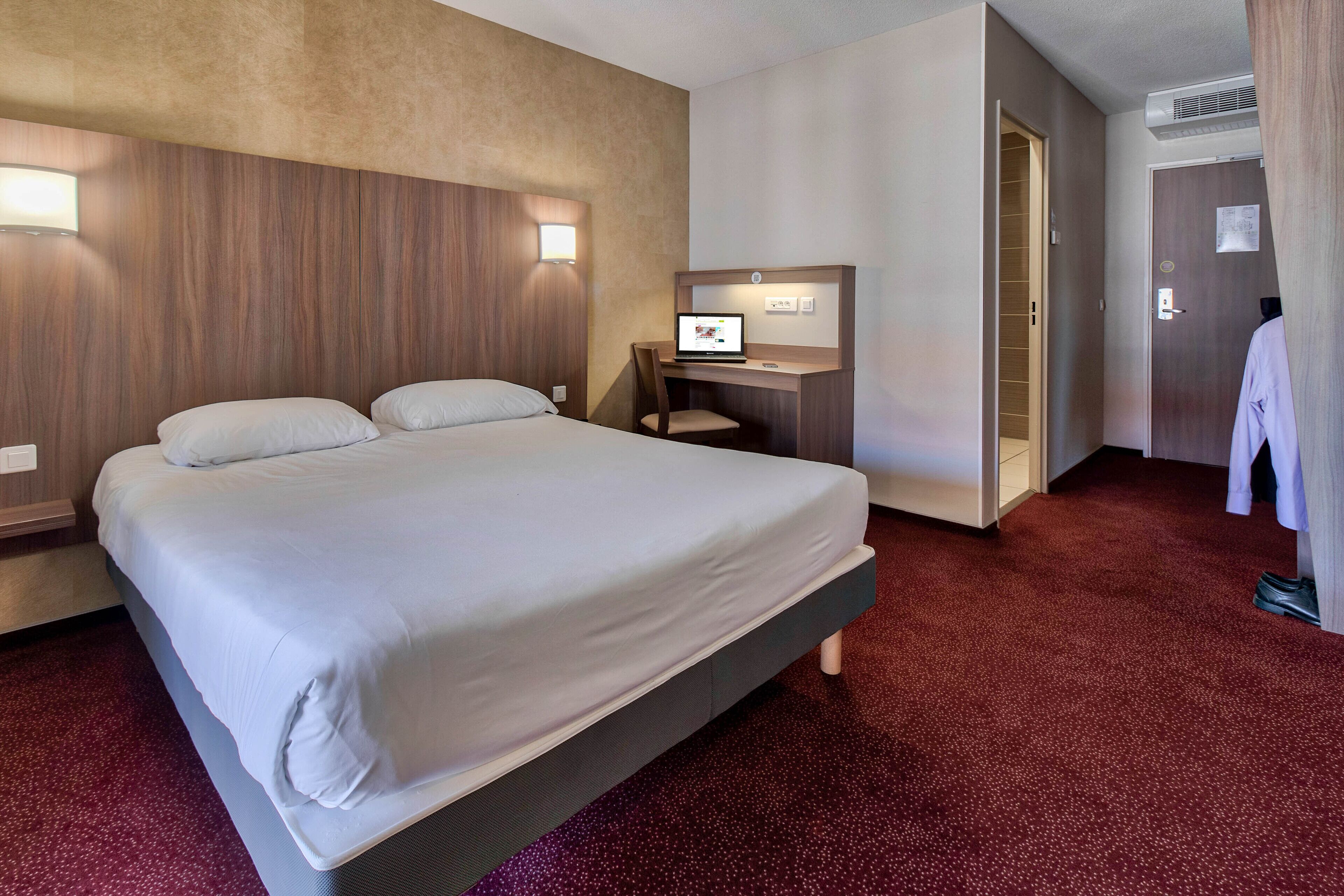 Et hotelværelse med en stor seng, et skrivebord med en bærbar computer og et knagered.