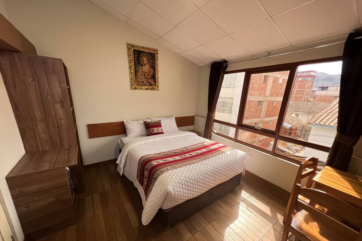 A hotel room with a wooden bed, a large window with a city view, and a wooden wardrobe.