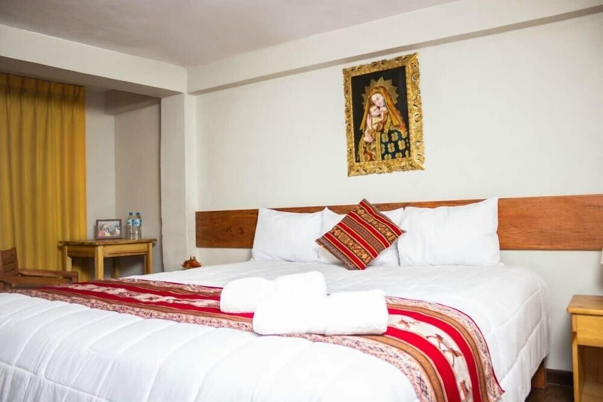 A hotel room with a neatly made bed, a wooden headboard, a wall-mounted religious painting, and a small bedside table with a bottle and a picture frame.