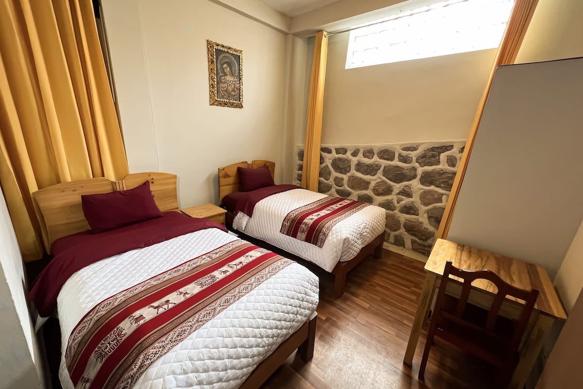 A room with two beds, wooden headboards, a stone wall, a small wooden table, and a chair.
