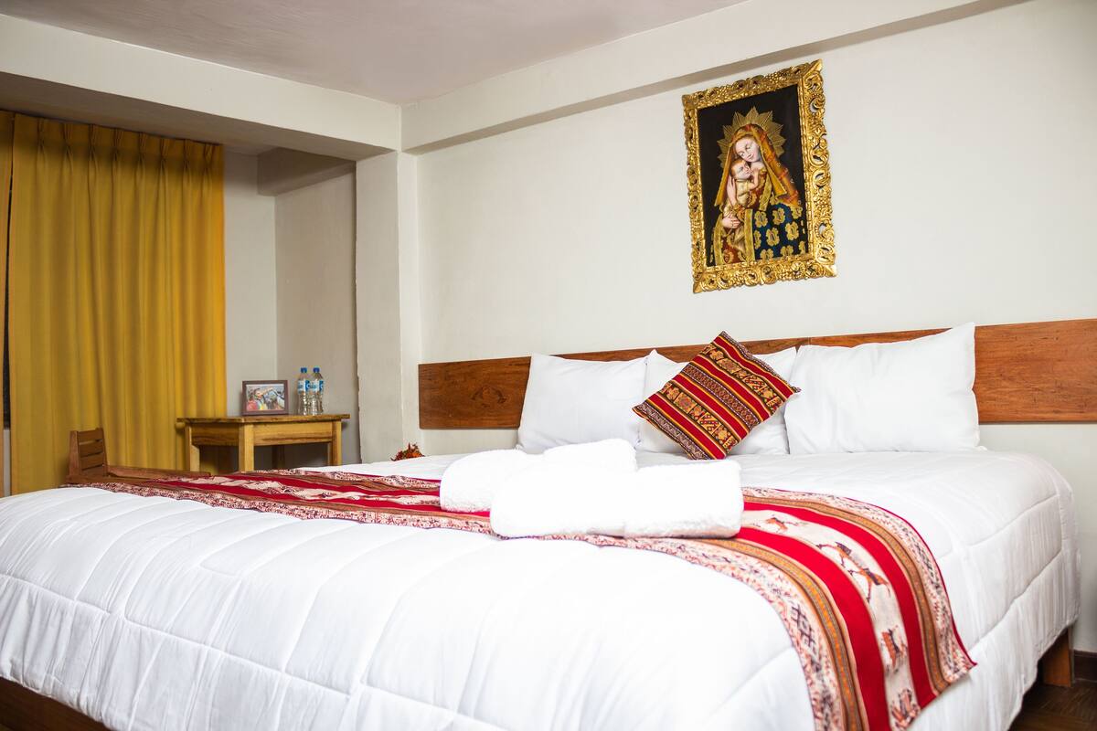 A hotel room with a neatly made bed, a wooden headboard, and a framed religious painting on the wall.